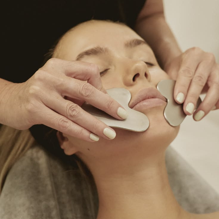 A close-up of a woman receiving a facial massage treatment with two silver metal gua sha tools on her face, held by a therapist wearing a black uniform.