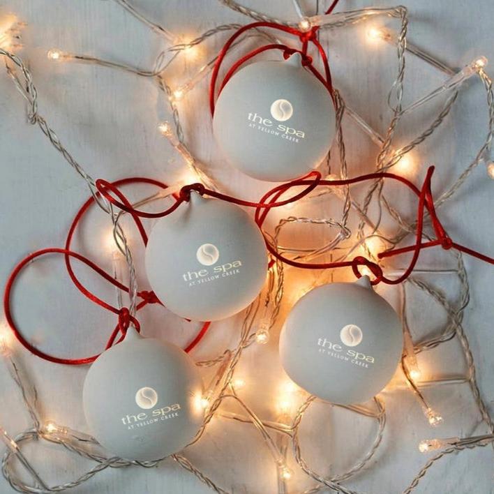 A luxury magnetic gift box tied with a satin ribbon, displaying the text 'The Spa' on multiple spherical gift items, surrounded by red and white Christmas lights.
