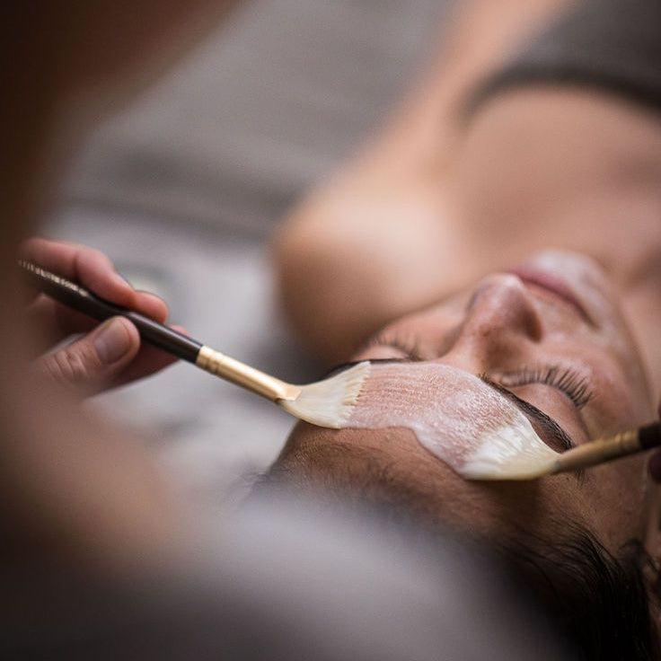 A close-up lifestyle shot of a person receiving a facial treatment, with a hand holding a makeup brush applying cream or mask to the cheek while another brush is nearby.