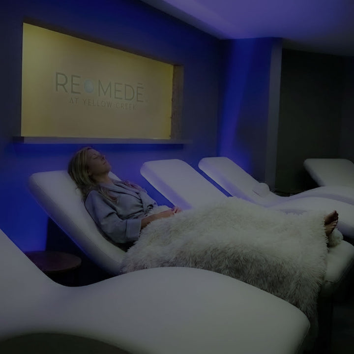 A woman relaxing on a white lounge chair in a dimly lit, modern spa relaxation room with blue ambient lighting at the Remede Lounge inside The Spa at Yellow Creek.