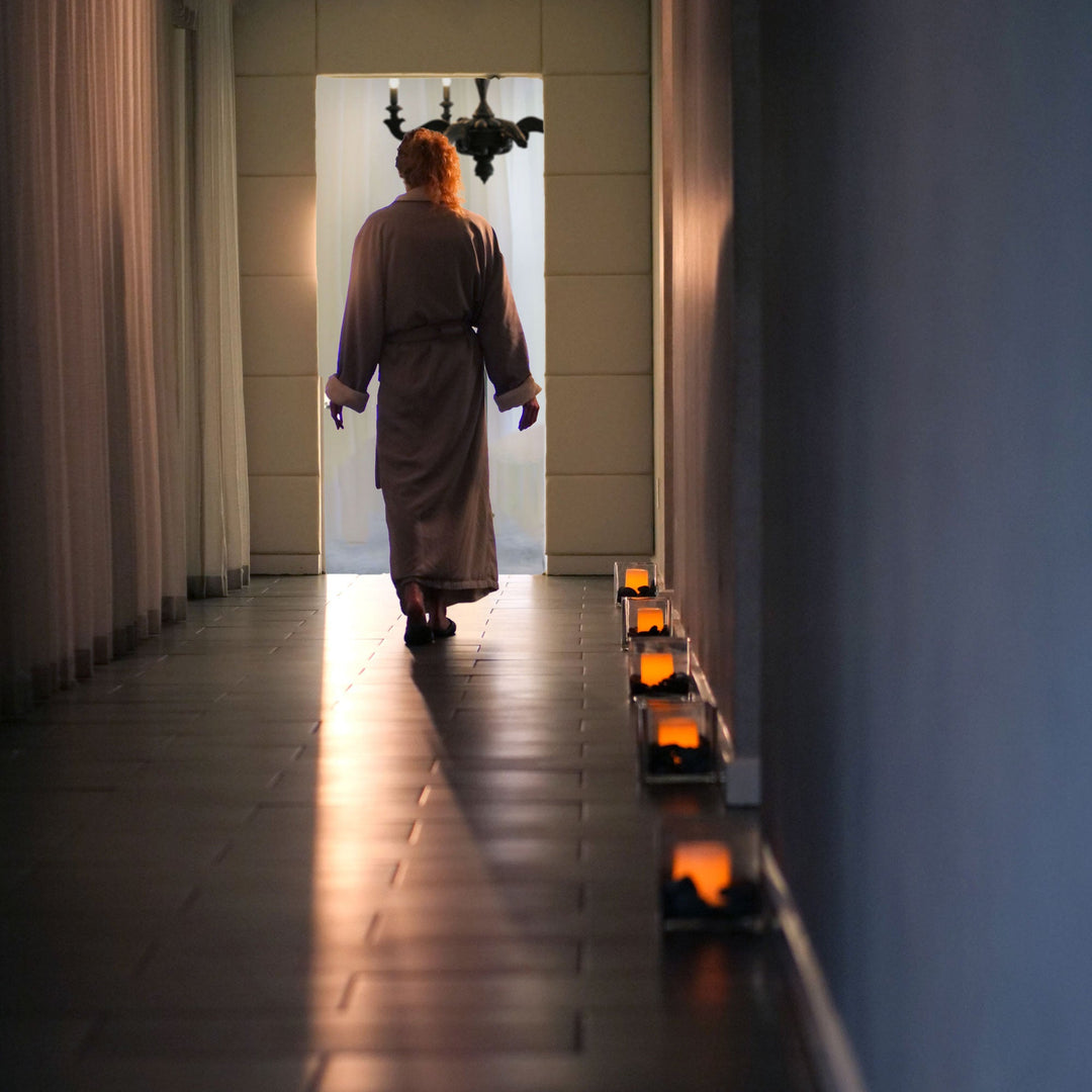 A person viewed from behind wearing a long taupe robe, walking down a dark tiled hallway toward a brightly lit doorway, with small glowing candles placed along the right wall.
