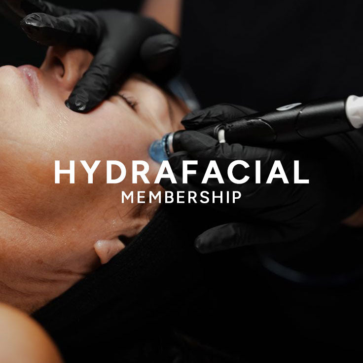 Close-up of a person receiving a hydrofacial treatment, with gloved hands holding the face open and applying a device to the skin. Large white text across the center reads 'HYDRAFACIAL MEMBERSHIP'.