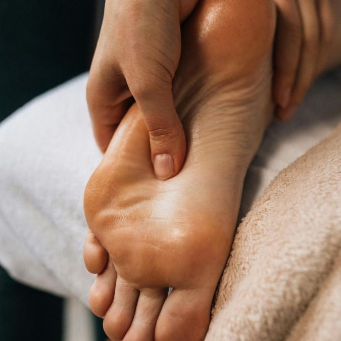 A close-up of a person's foot being massaged by another person's hands, with a soft beige towel visible in the foreground.